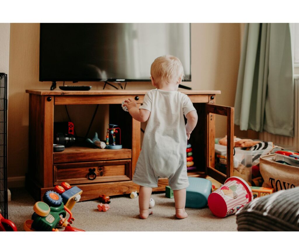 a baby standing amidst many toys in a living room mess.